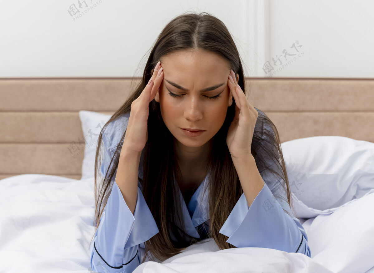 美丽穿着蓝色睡衣的年轻美女躺在床上抚摸着太阳穴 在明亮的背景下感受卧室内部的疼痛痛苦蓝色睡衣