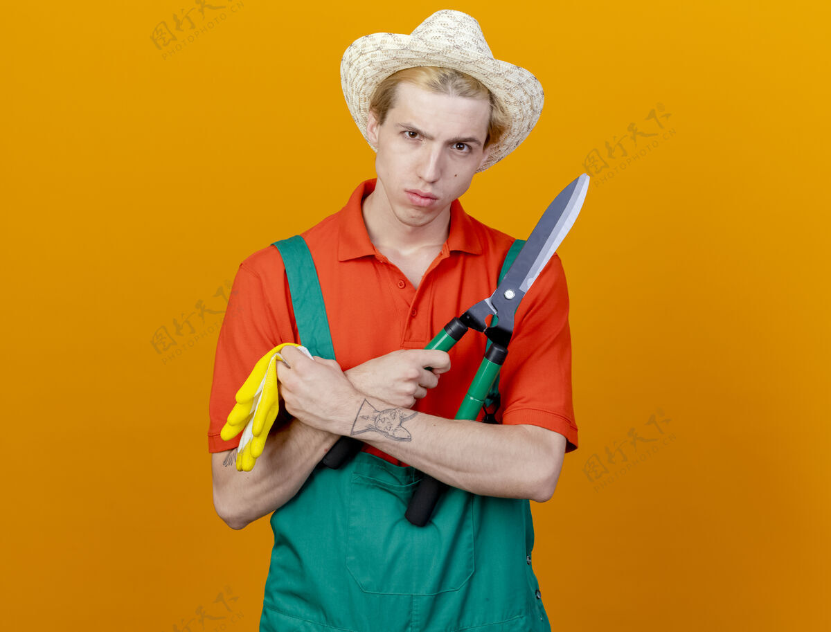篱笆年轻的园丁穿着连体衣 戴着帽子 拿着树篱剪和橡胶手套 严肃的脸站在橙色背景上看着摄像机橙色穿园丁