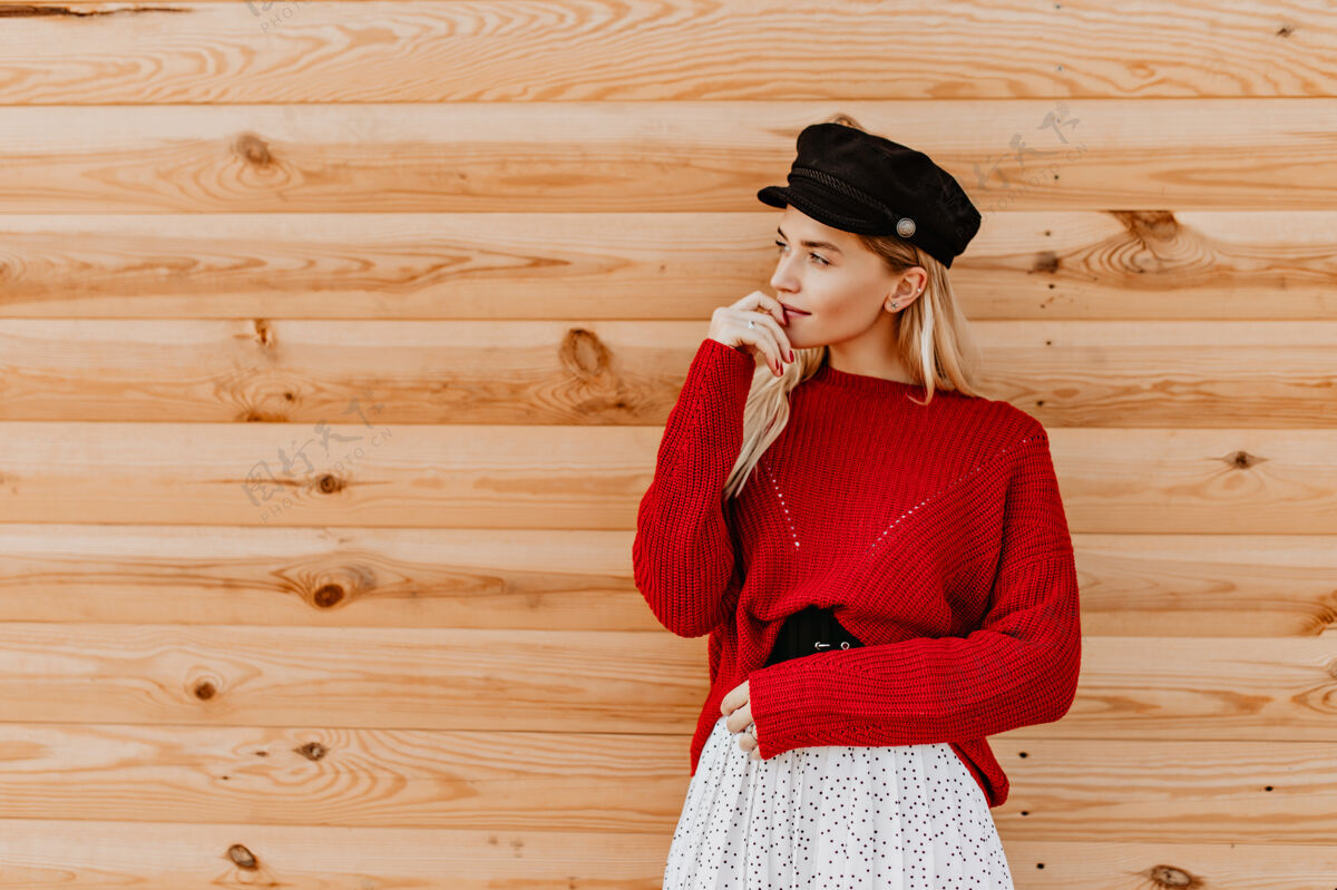 房子美丽的金发女郎 戴着黑帽子 穿着漂亮的红毛衣 在木墙上温柔地摆着姿势迷人的女孩在外面享受着温暖的秋日光木头户外