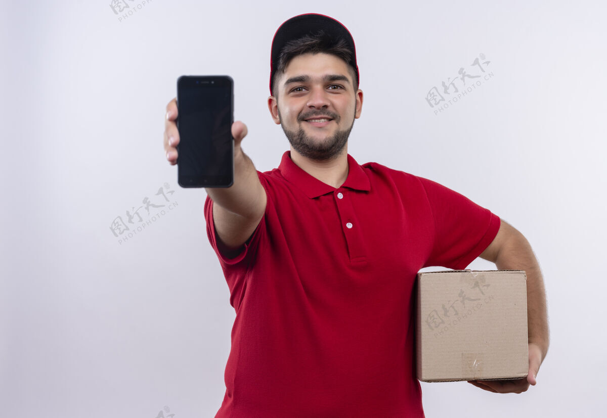 拿着年轻的送货员穿着红色制服 戴着帽子拿着盒子包 展示着智能手机 自信地微笑着看着相机帽子制服男人