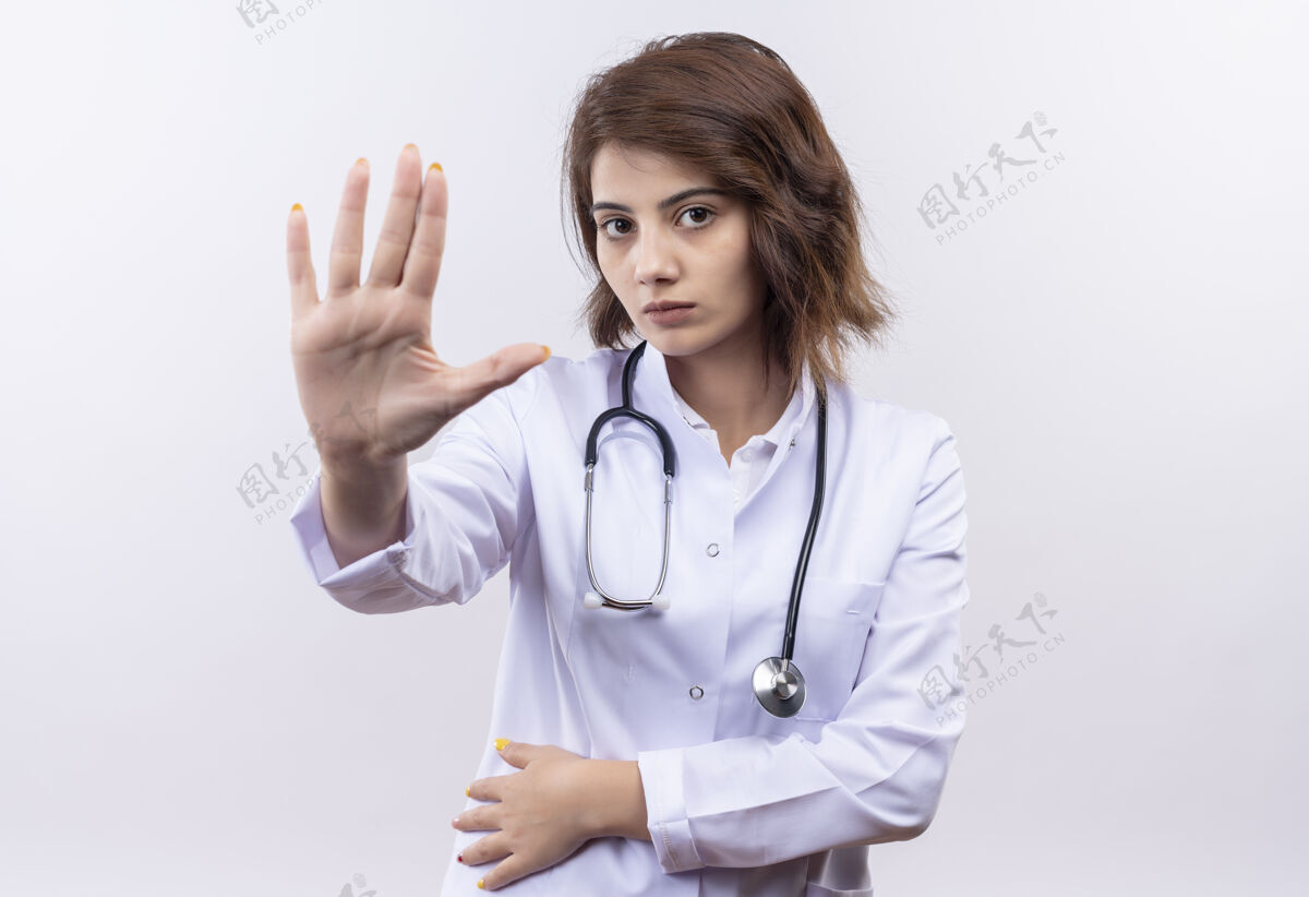 女人身穿白大褂 手持听诊器的年轻女医生站在那里 双手张开 打着停车牌 严肃地看着镜头 背景是白色的认真听诊器做