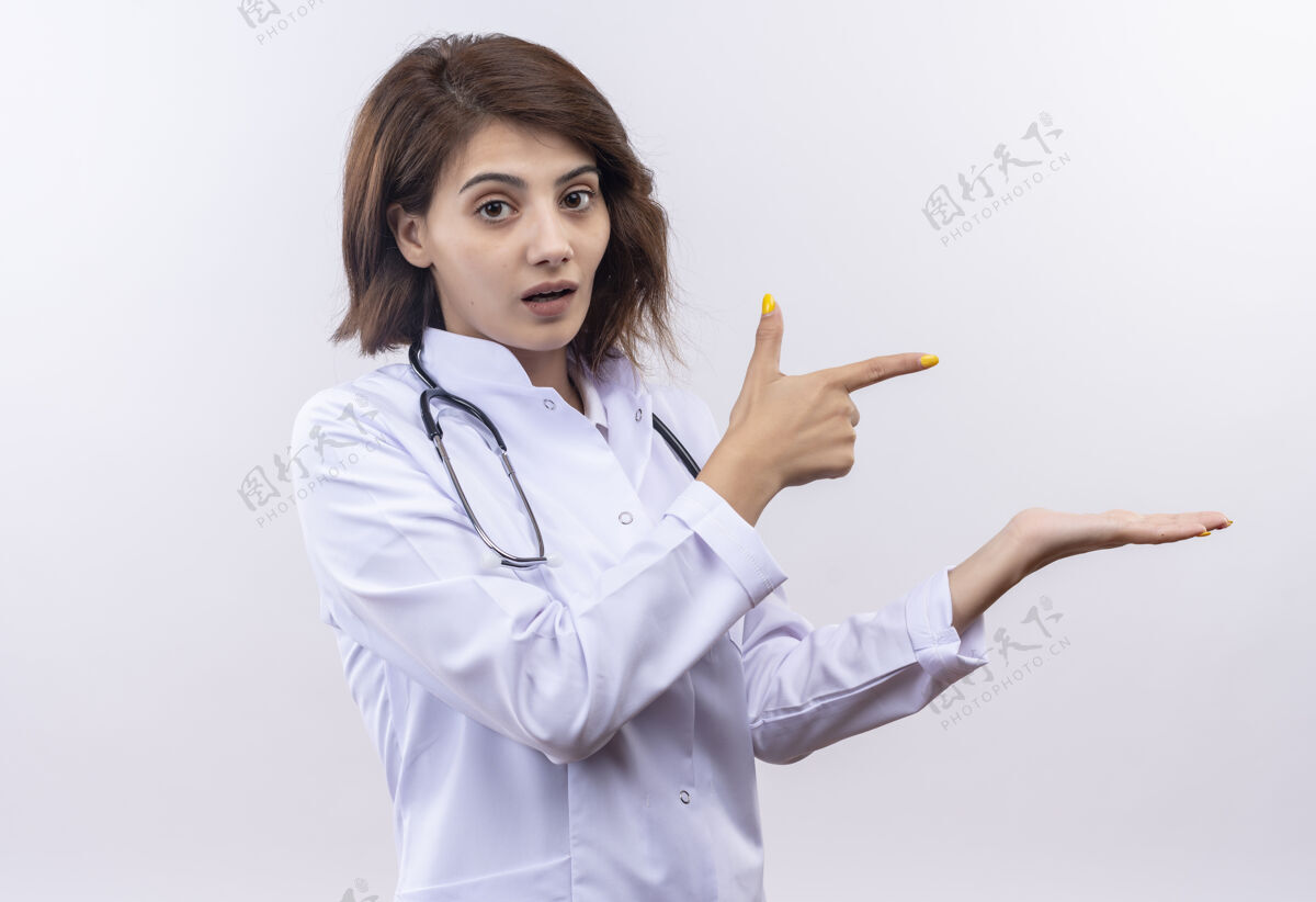 食指身穿白大褂 手持听诊器的年轻女医生自信地用食指指着侧面看站着医生