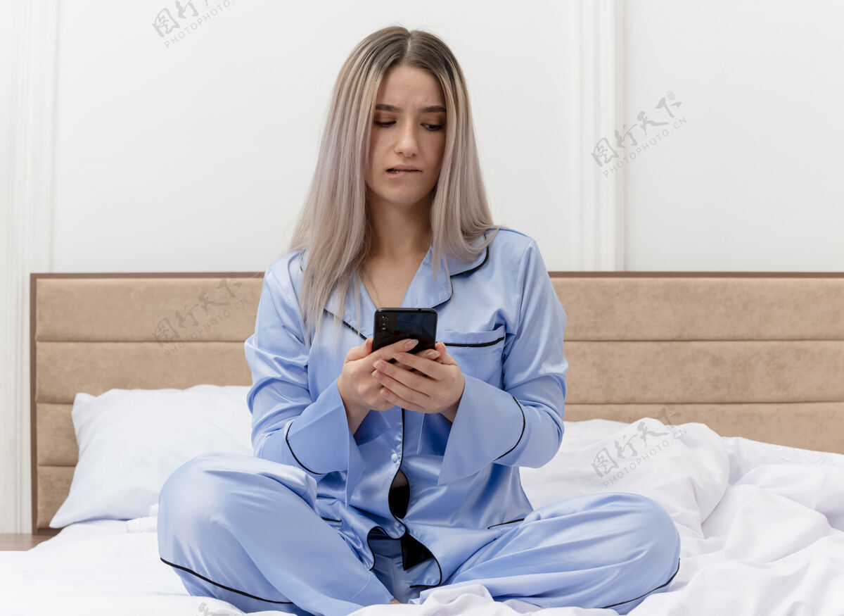 看穿着蓝色睡衣的年轻美女坐在床上用智能手机看着困惑的咬着嘴唇在卧室的室内灯光背景下嘴唇困惑咬