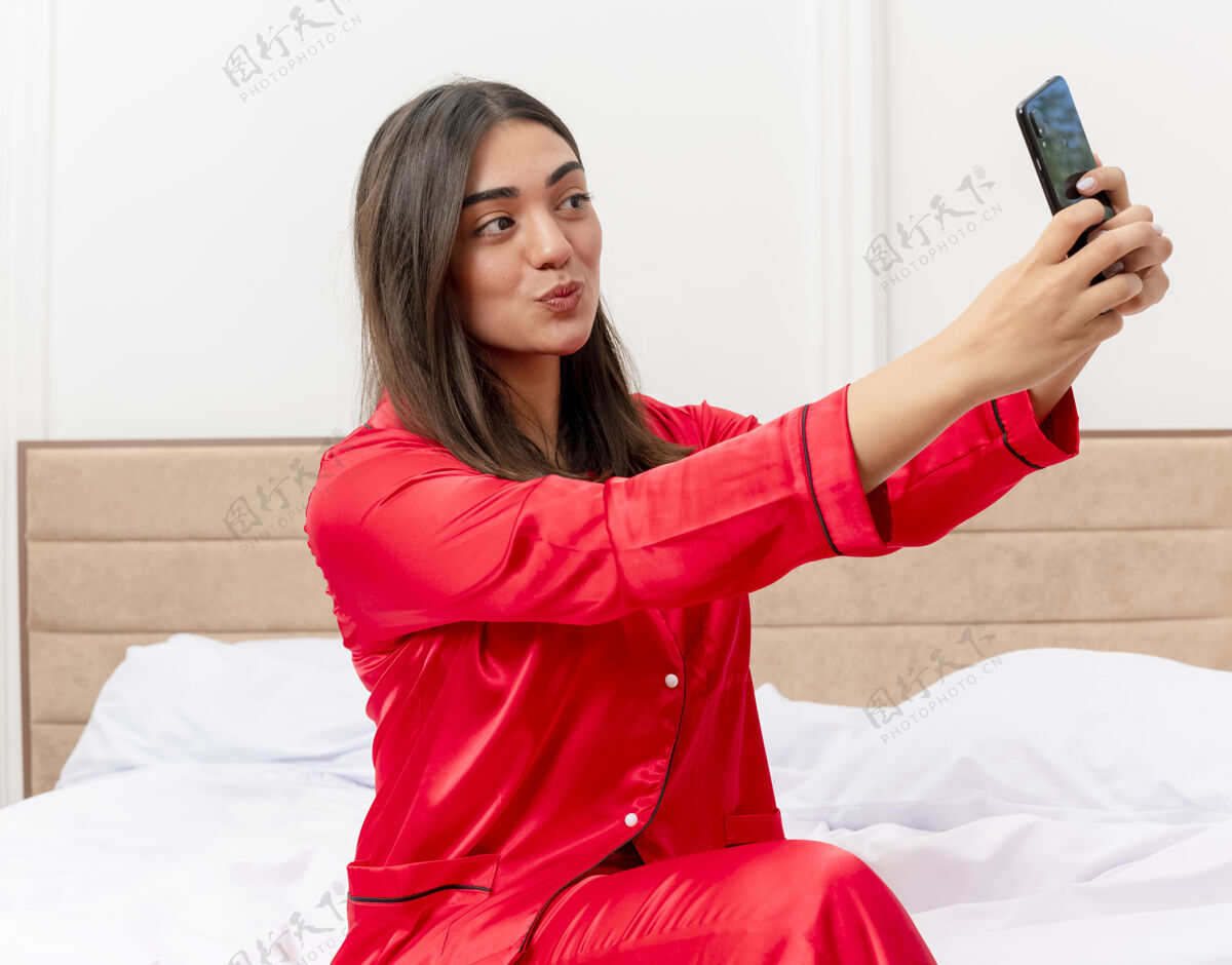 漂亮穿着红色睡衣的年轻漂亮女人坐在床上用智能手机自拍 在卧室的室内灯光背景下开心而积极地微笑自拍微笑睡衣