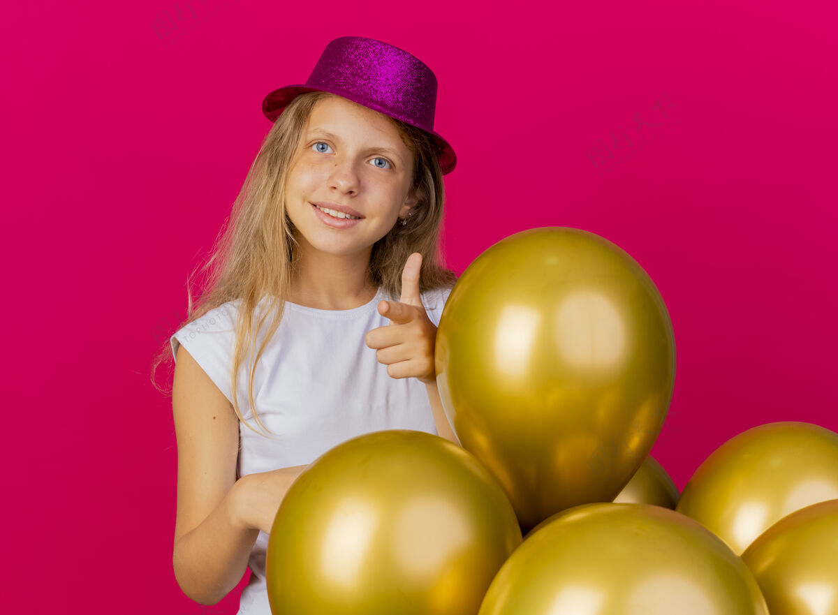 高兴戴着节日礼帽的快乐可爱的小女孩 一群巴隆人微笑着用食指指着镜头 粉色背景上站着生日派对的概念束站着指着