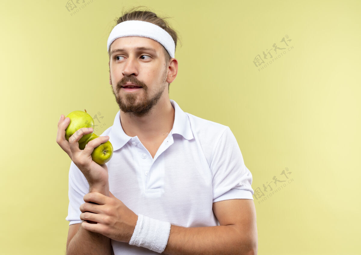 帅气体贴的年轻帅气的运动型男人戴着头带和腕带拿着苹果 握着手腕望着绿色空间里与世隔绝的一面手腕穿复制