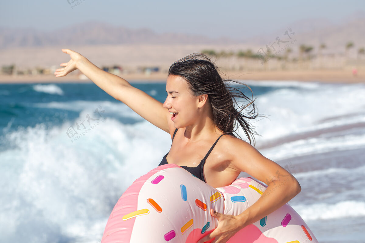 夏天一个快乐的年轻女子 在海边有一个甜甜圈形状的游泳圈度假休闲娱乐的概念黑发晒黑女人