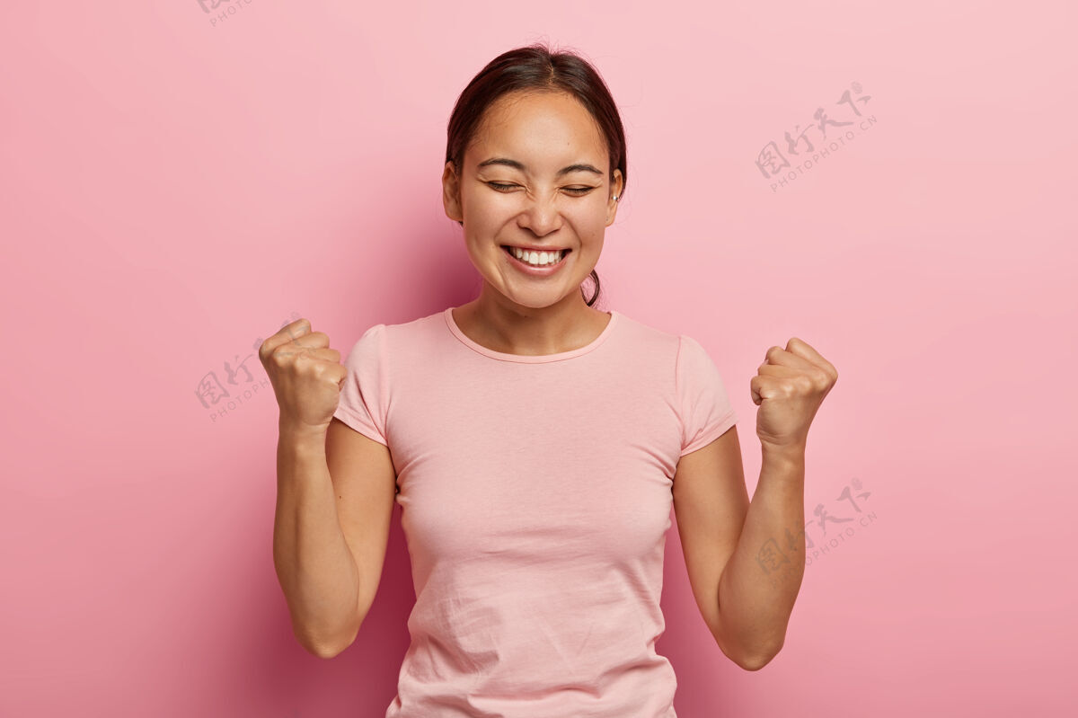 女士成功感性的亚洲女人在空中握紧拳头 庆祝成功 笑容灿烂 闭上眼睛 兴奋而快乐 享受胜利的甜味 穿着休闲服提高微笑喜悦