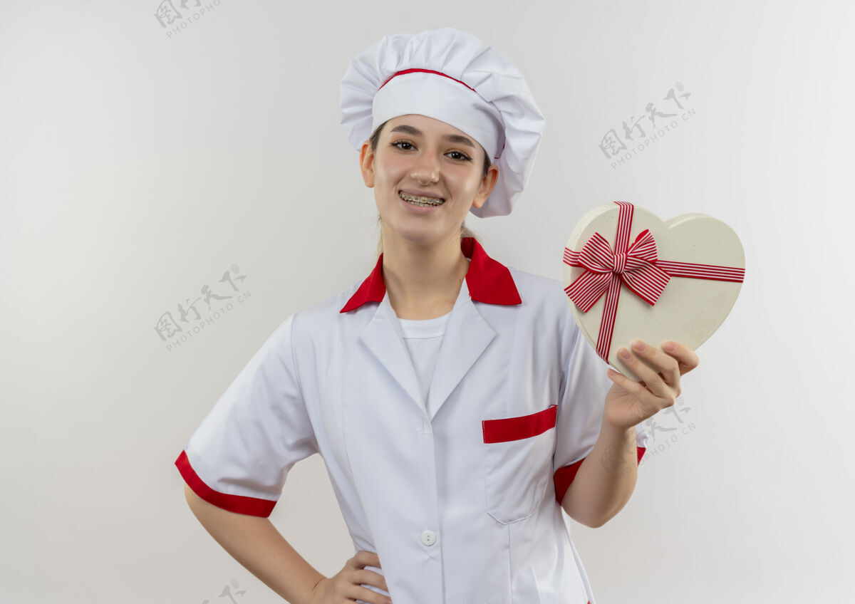 牙科微笑的年轻漂亮厨师 身着厨师制服 戴着牙套 手放在腰上 手持心形礼品盒 与白色空间隔离年轻拿着微笑