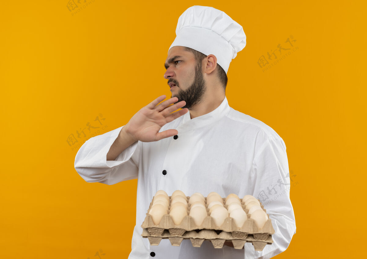 持有穿着厨师制服的年轻男厨师手拿着一盒鸡蛋望着旁边 在橙色的空间里没有孤立的手势橙色男性未发布