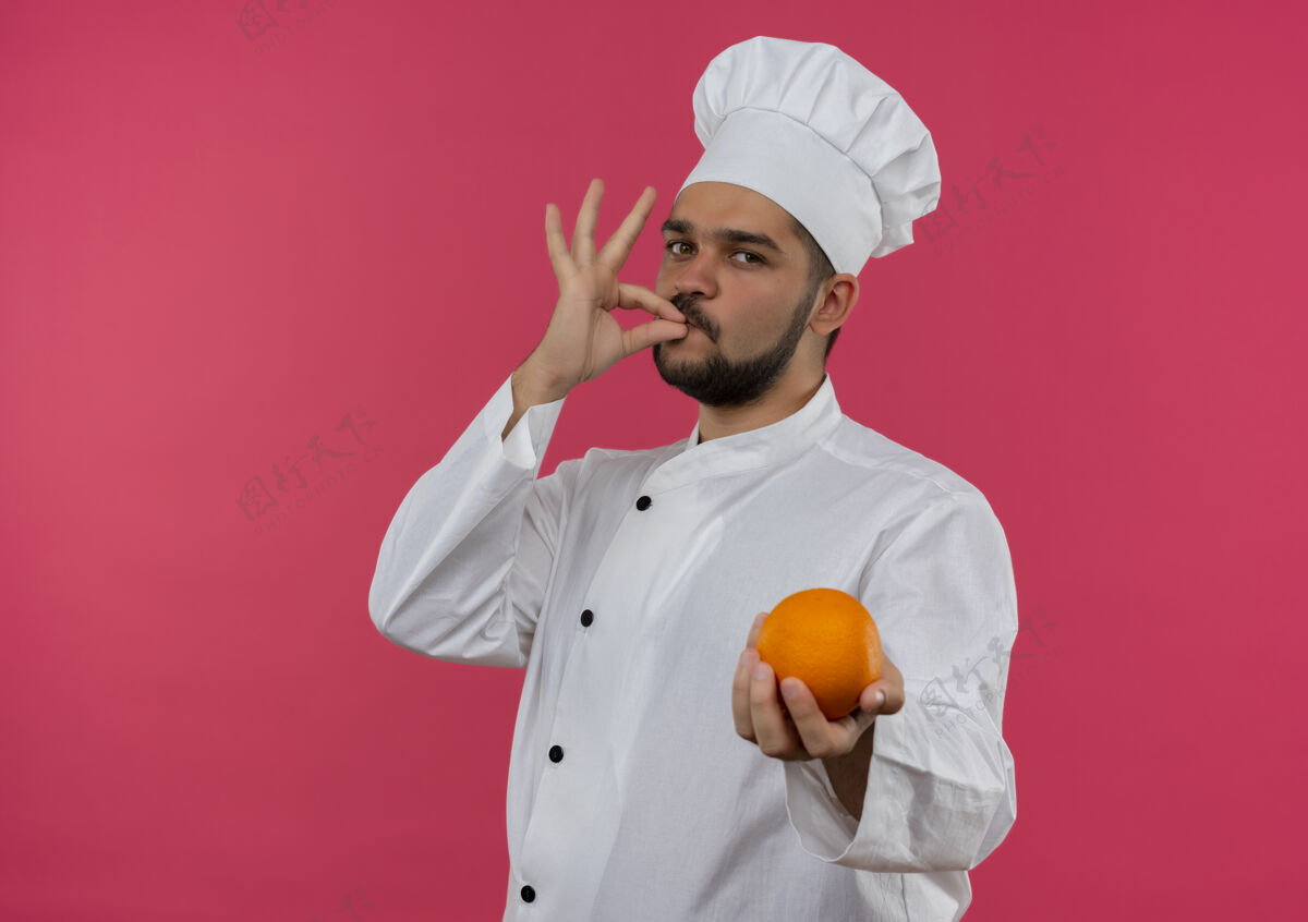 做身着厨师制服的年轻男厨师手拿橘子 做着美味的手势 看上去孤立在粉色的空间里手势看制服