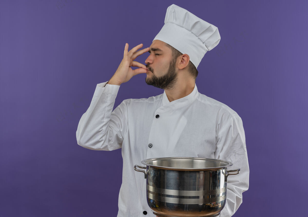 美味年轻的男厨师穿着厨师制服 拿着锅 做着美味的手势孤立在紫色的空间里男性做制服