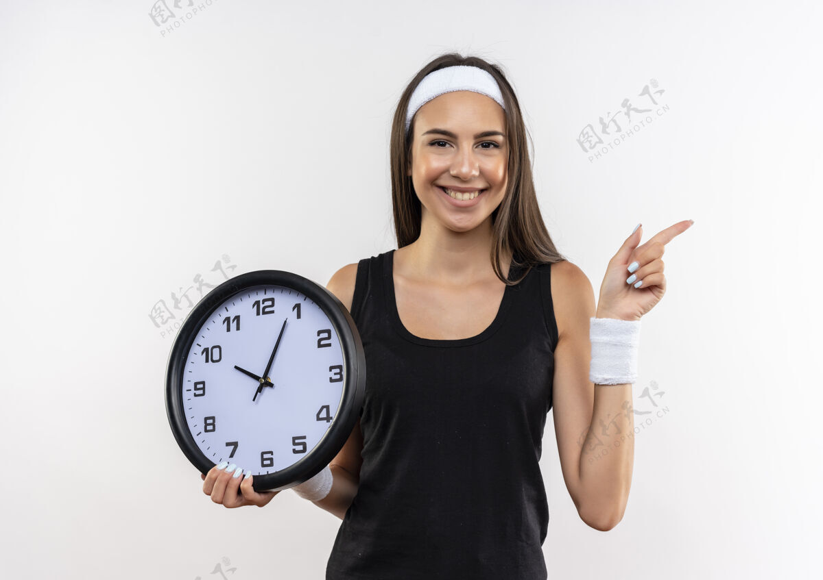 腕带微笑着漂亮的运动女孩戴着头带和腕带拿着时钟 指着隔离在白色空间的一边女孩抱着时钟