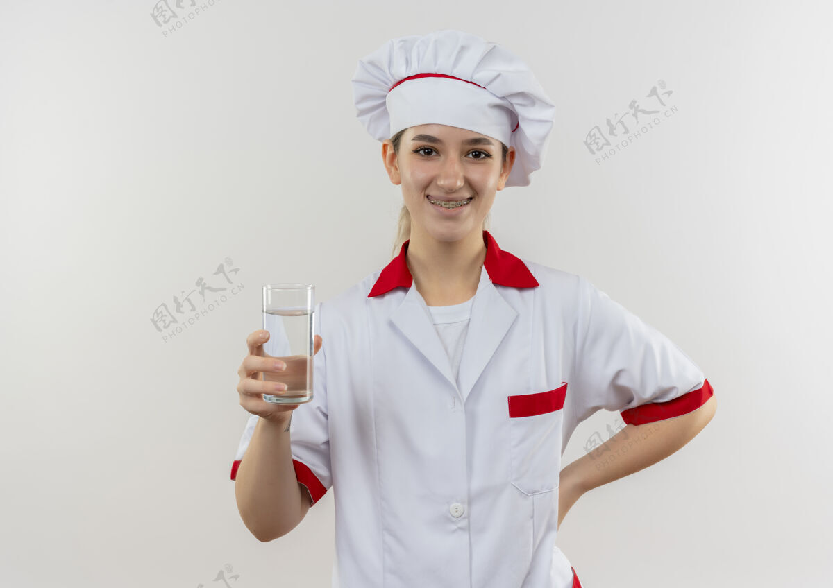 漂亮面带微笑的年轻漂亮厨师 身着厨师制服 戴着牙套 端着一杯水 把手放在腰部 与白色空间隔离开来微笑厨师水