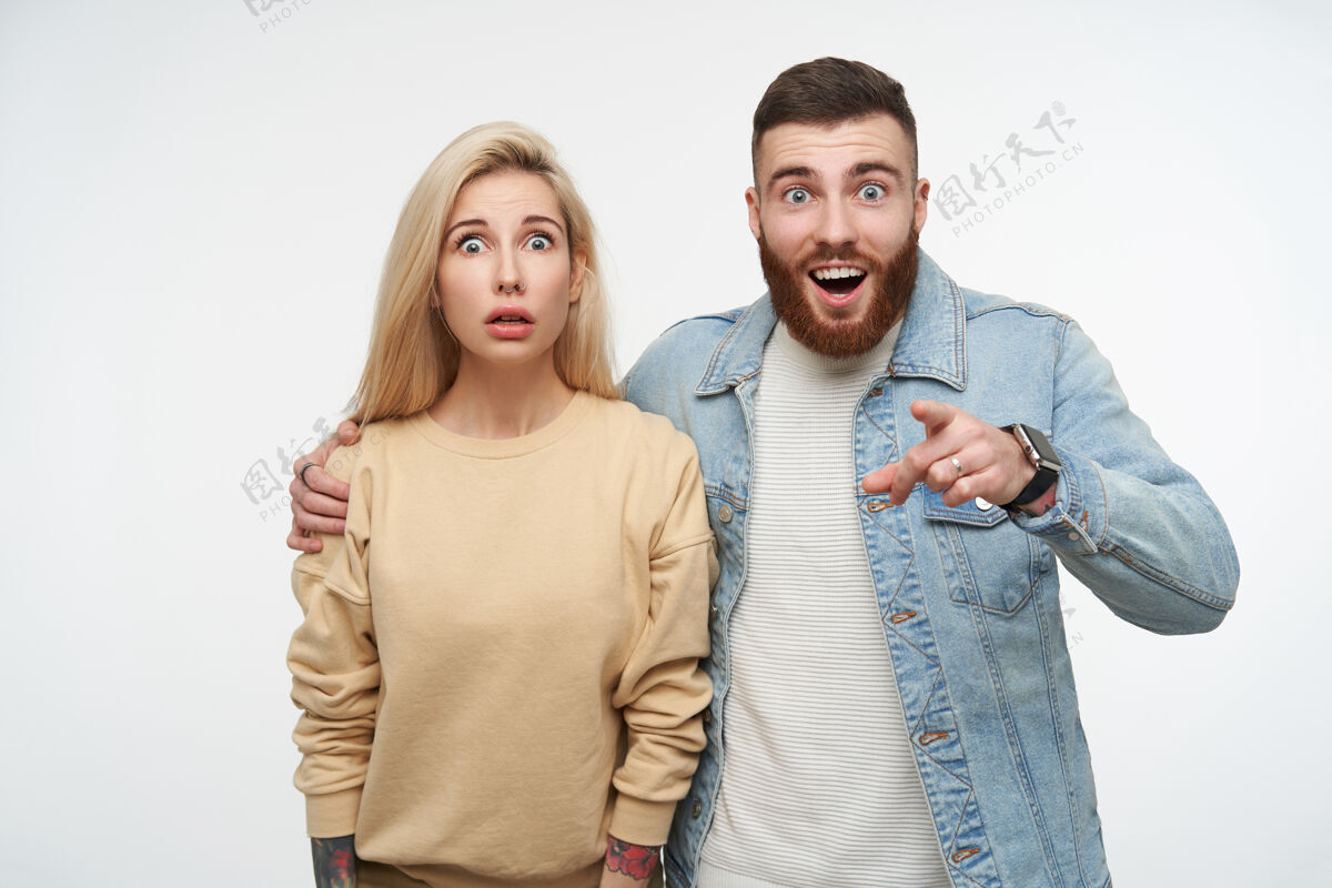 站立一对年轻的蓝眼睛漂亮夫妇的室内肖像 穿着米色运动衫和牛仔裤外套 站在白色的地面上 脸上带着震惊的表情年轻情感双人