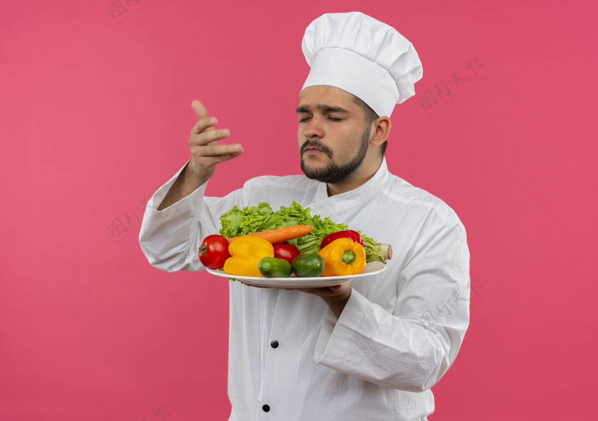 厨师身着厨师制服的年轻男厨师手拿着一盘蔬菜 闭着眼睛嗅着 被隔离在粉红色的空间里盘子粉红色嗅