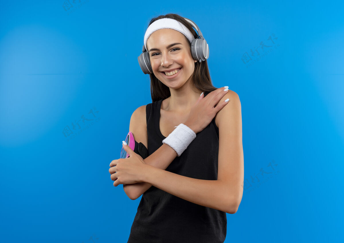 电话微笑的年轻漂亮的运动女孩戴着头带和腕带和耳机与手机袖标把手放在肩膀隔离在蓝色空间腕带运动女孩