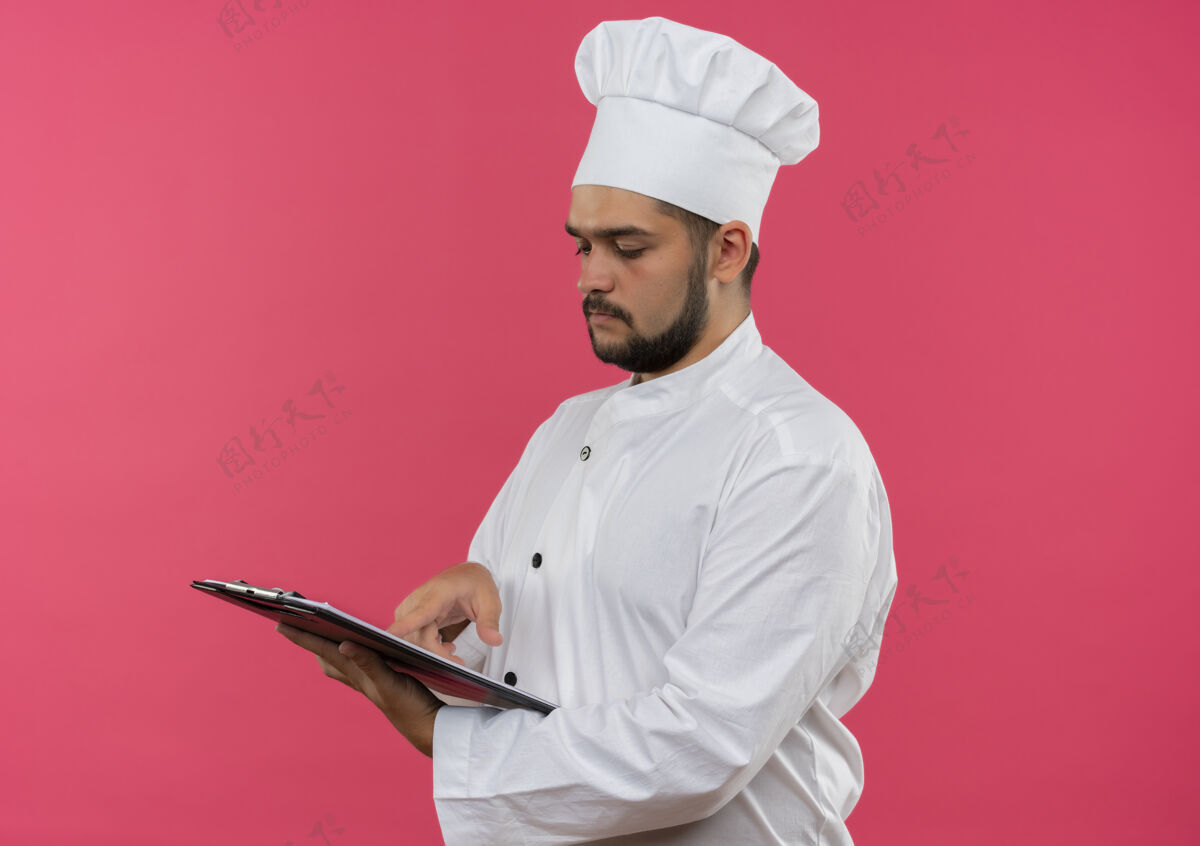 年轻年轻的男厨师穿着厨师制服 拿着看 把手指放在剪贴板上孤立的粉红色空间剪贴板男空间