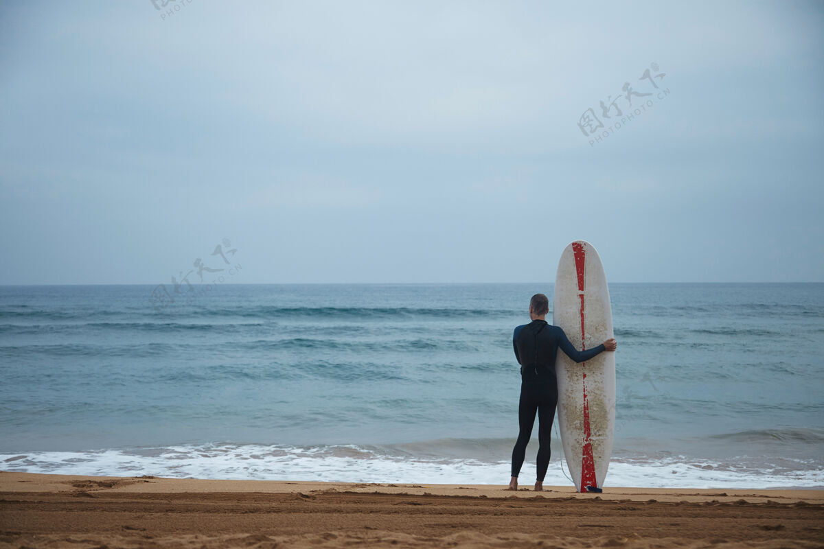 度假带着长冲浪板的老冲浪者一个人呆在海边看海浪 然后去冲浪 一大早穿着全套的潜水衣太阳视图热带