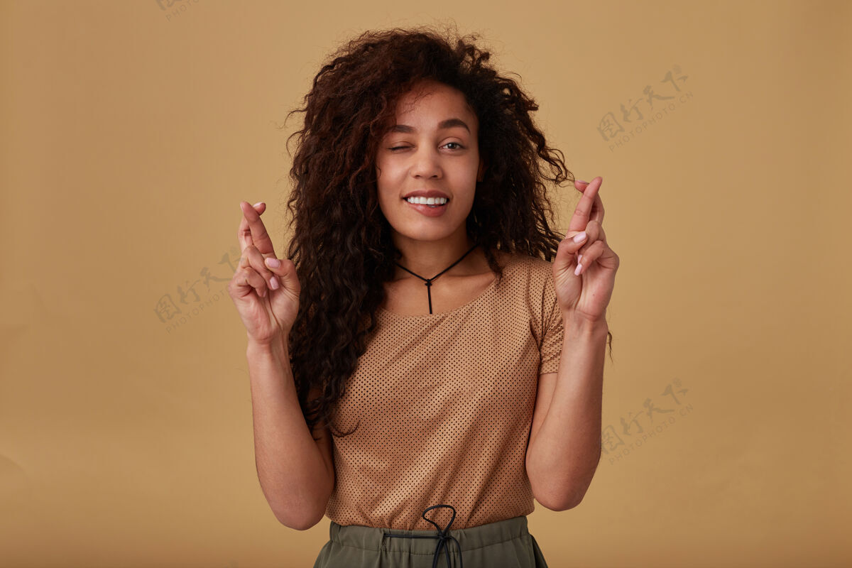 米色室内肖像的年轻积极卷曲黑皮肤黑发女士举手交叉手指和咬令人不安的臀部 而在米色摆姿势交叉情绪站立