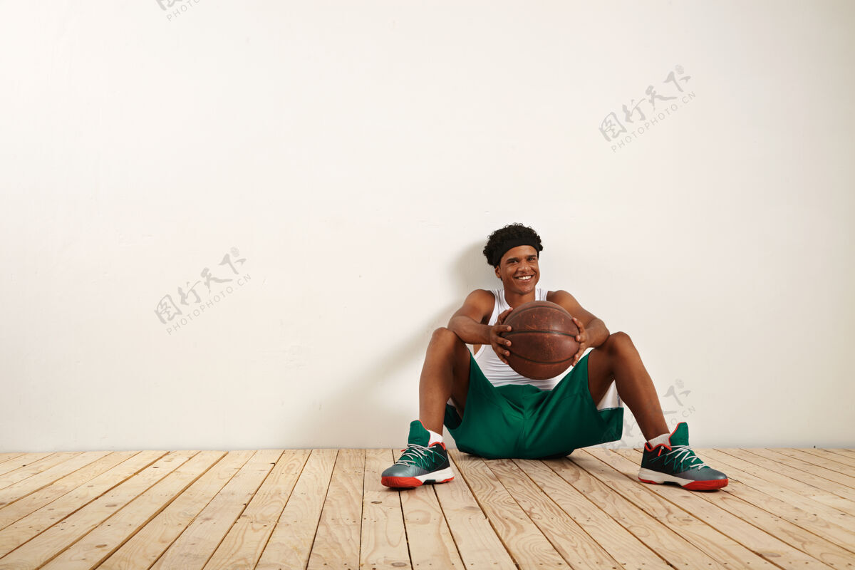 篮球一个微笑的年轻球员坐在木地板上 靠着一堵白色的墙 手里拿着一个棕色的旧篮球成人服装团队