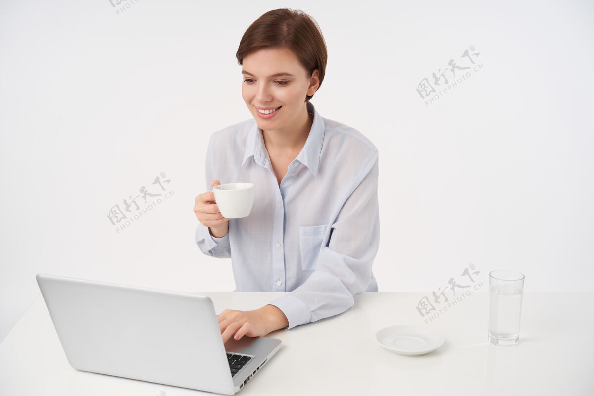 水可爱的年轻漂亮的棕色头发的女士 留着时髦的短发 一边看邮箱一边积极地微笑 一边摆着白色的姿势喝着茶模特情绪摆姿势