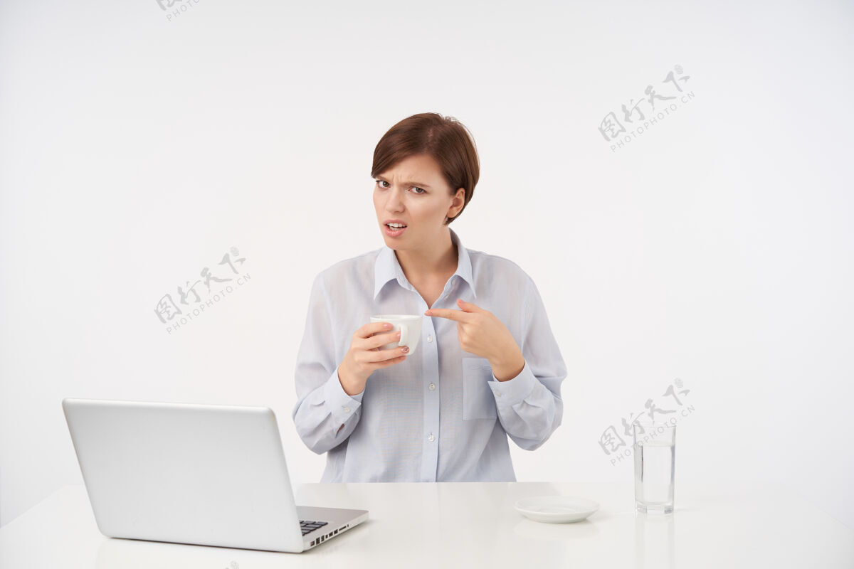 短不满意的年轻漂亮的棕色头发的女士 短发时髦 手里拿着陶瓷杯 用食指指着它 坐在白色的椅子上困惑地看着坐着优雅员工