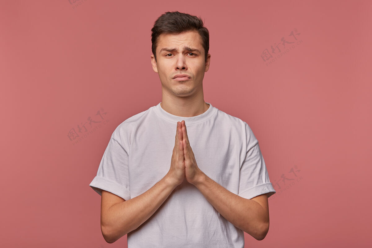 不快乐一个穿着白t恤的年轻人 希望好运 做着祈祷的手势 站在粉红色的地面上 表情很不高兴怨恨英俊悲伤