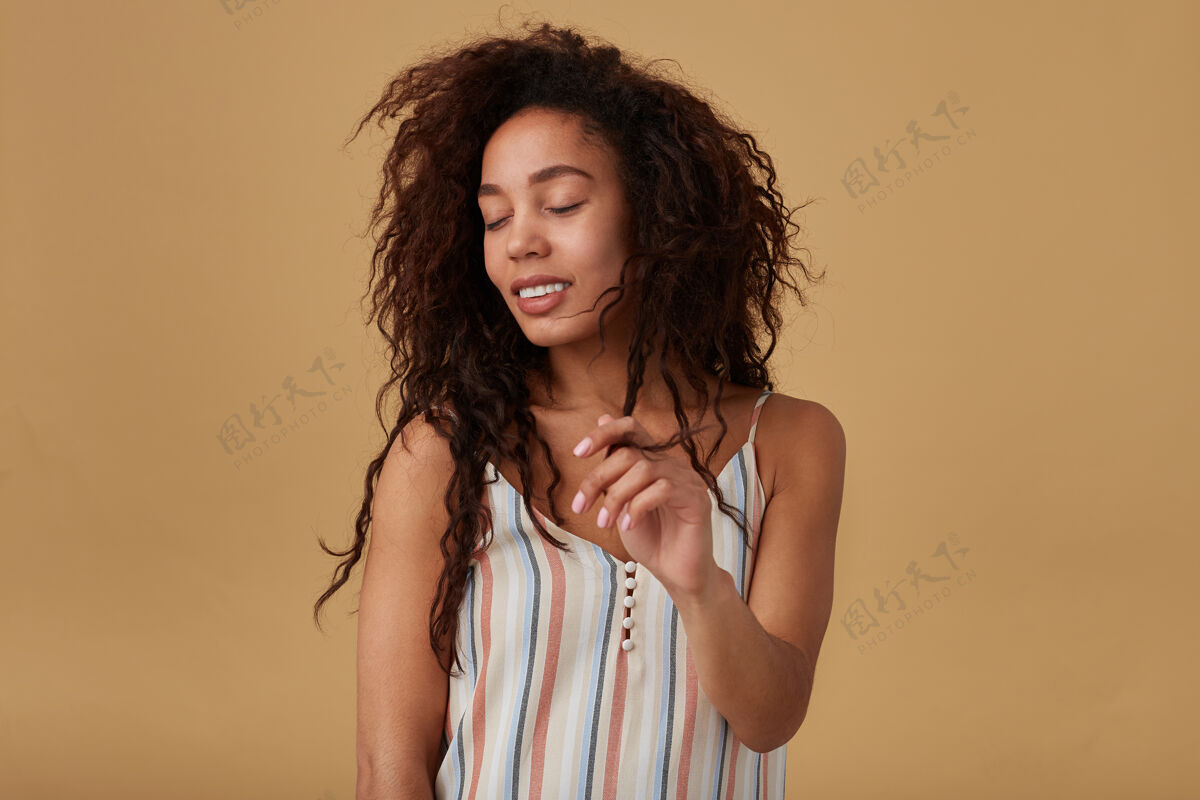 高兴年轻漂亮的棕色头发卷曲的深色皮肤女士 自然的妆容 双手举起 面带微笑 摆出米色的姿势卷发休闲摩卡