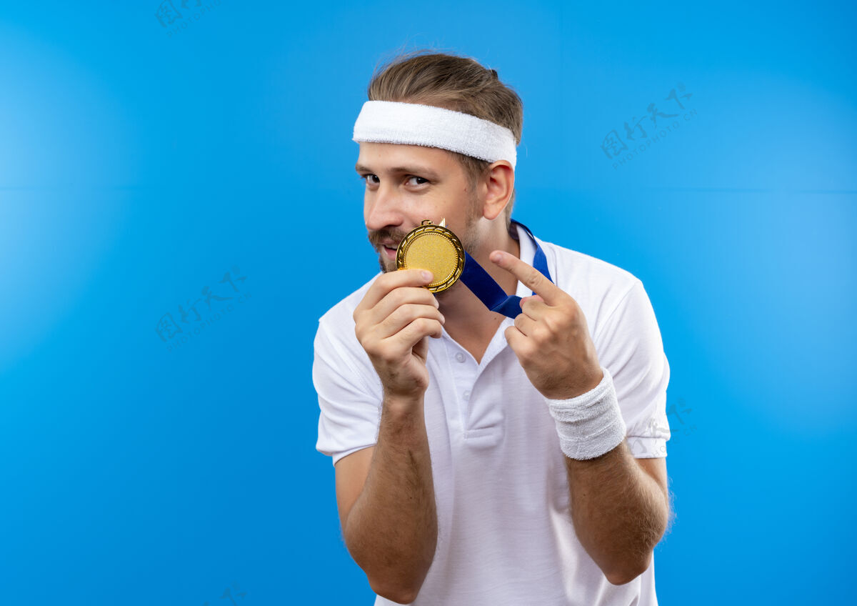 男人高兴的年轻帅气的运动型男子戴着头带和腕带 围着奖牌的脖子拿着和指着奖牌孤立的蓝色空间拿着帅气蓝色