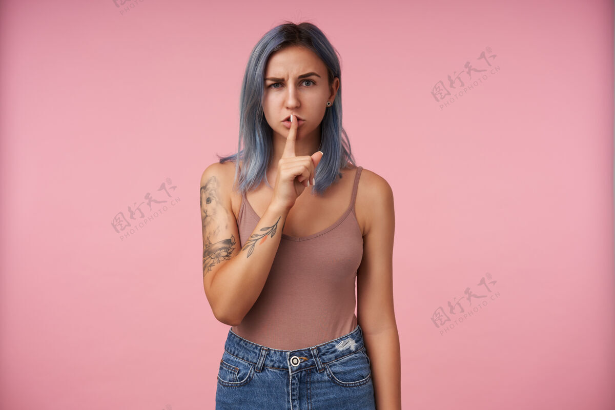 蓝色室内肖像年轻严重纹身女性保持食指在她的嘴唇上 而认真地看 穿着米色衬衫 而在粉红色的姿势休闲室内纹身