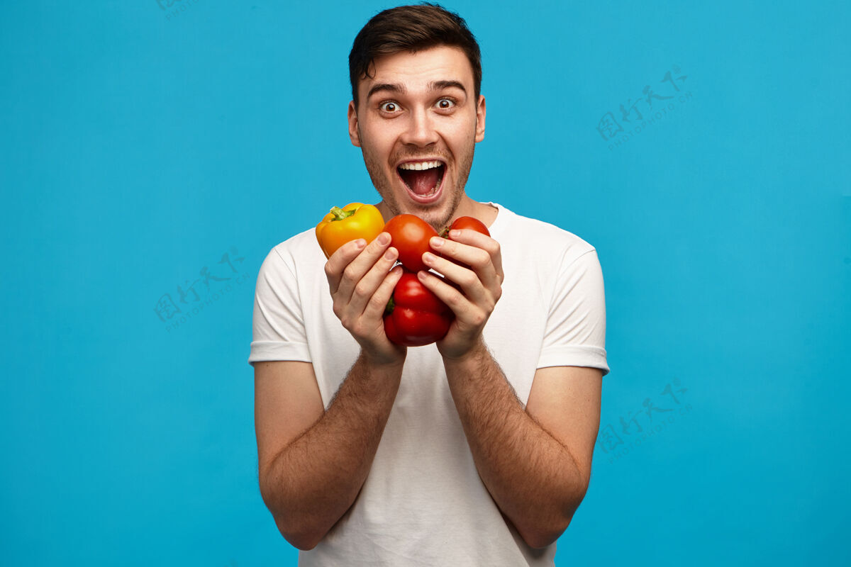 年轻一个穿着白衬衫 情绪激动的年轻男子 两手拿着甜椒和西红柿 神情激动 张大嘴巴 满心欢喜地吃着花园里新鲜的有机蔬菜新鲜表情英俊