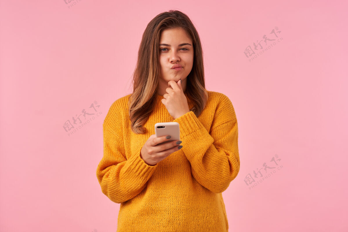 女性沉思的年轻棕色头发的女人 自然化妆 举起手托着下巴 眯着眼睛若有所思地撅着嘴唇 孤立在粉红色的脸上毛衣休闲运动衫