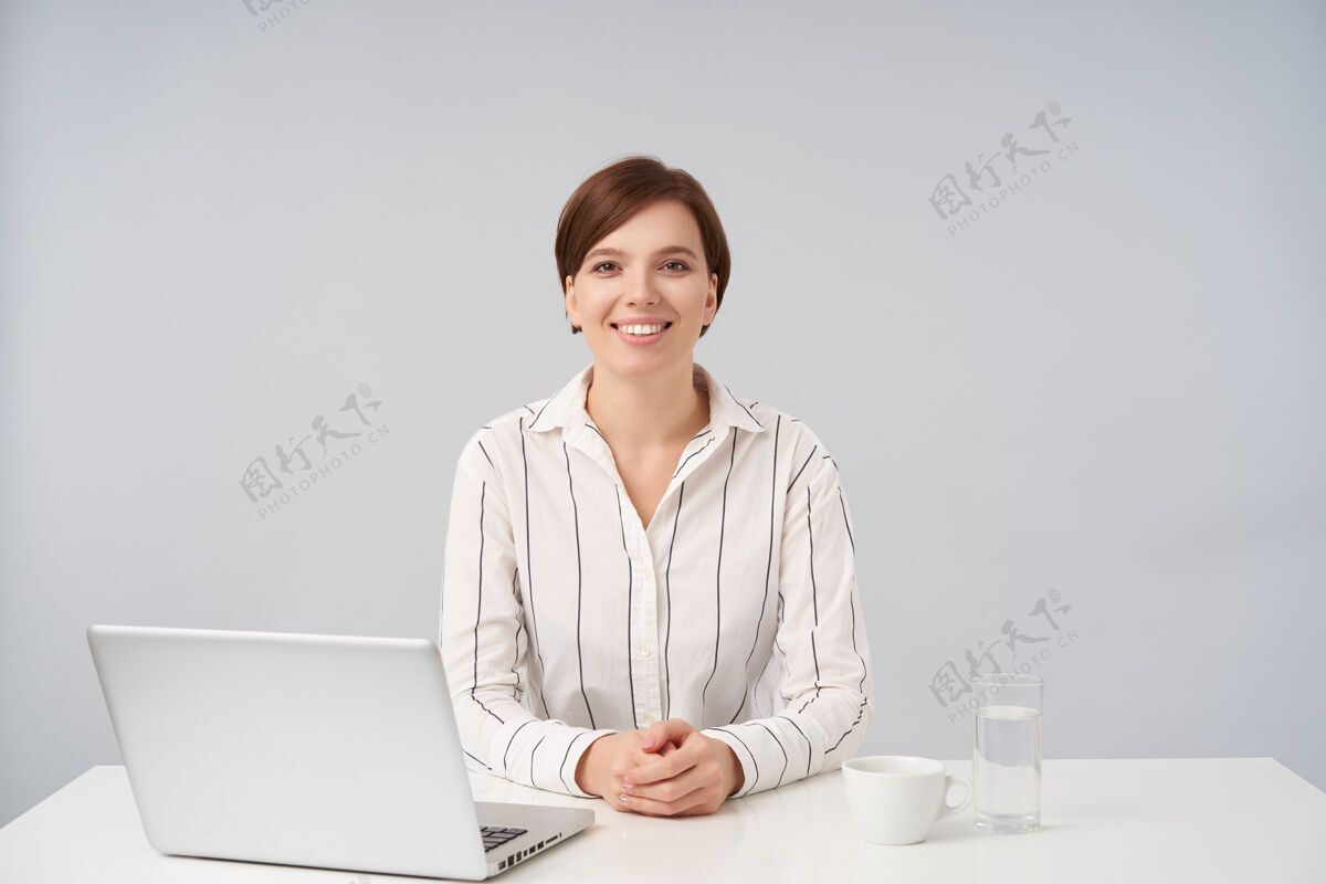 化妆品快乐的年轻棕色眼睛迷人的短发黑发女士保持她的手在台面上 而坐在桌子上的白色 面带笑容愉快地看着笔记本电脑短摆姿势
