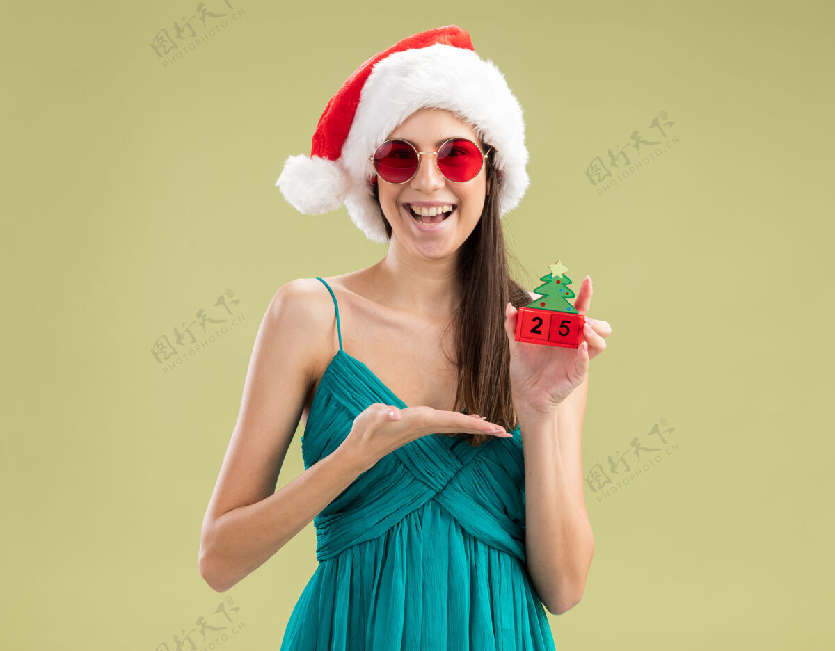复制戴着太阳眼镜 戴着圣诞帽 手拿圣诞树饰物的快乐白人女孩橄榄指向绿色