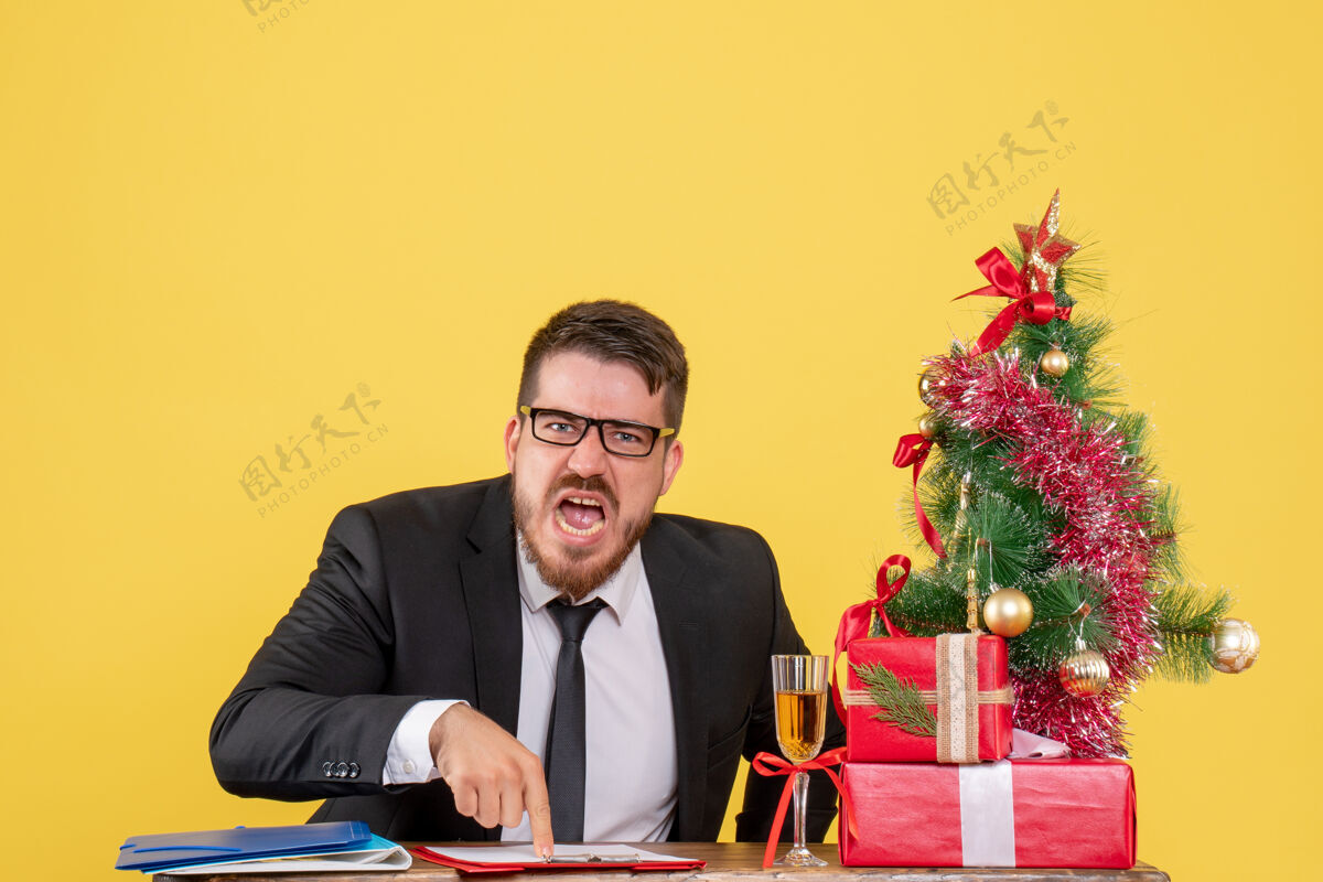 新郎正面图：桌子后面的男工人拿着礼物 圣诞树在黄色上尖叫商人工人圣诞