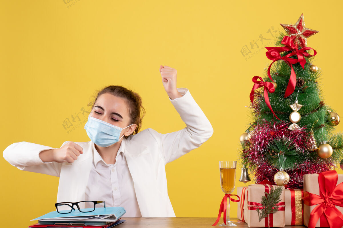 情绪正面图：女医生戴着无菌口罩坐在黄色背景上 带着圣诞树和礼品盒 欢呼雀跃衣服人实验室外套