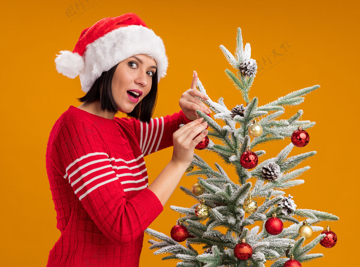 站着令人印象深刻的年轻女孩戴着圣诞帽站在侧视图附近的圣诞树装饰圣诞饰品看相机孤立在橙色背景装饰个人资料年轻