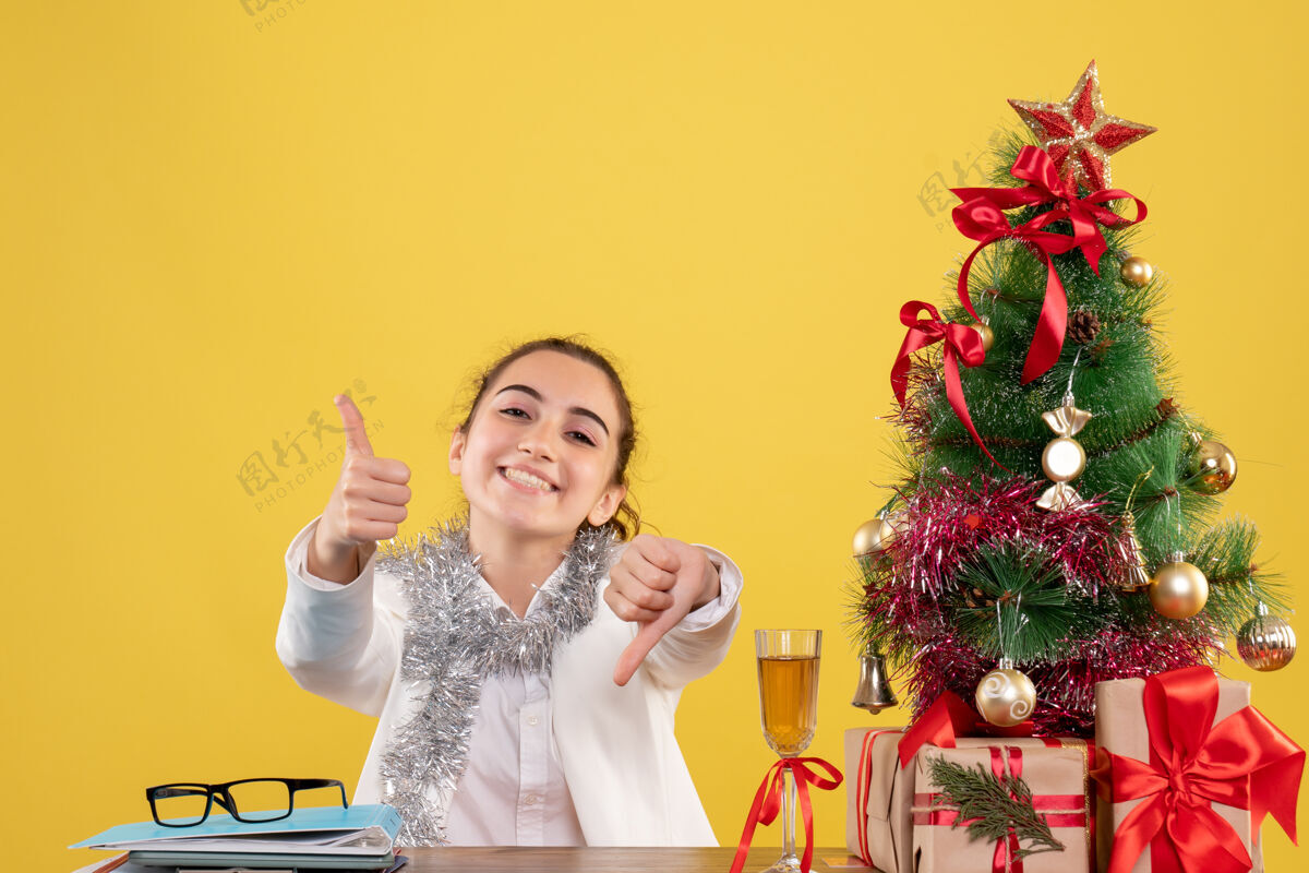 快乐正面图：女医生坐在黄色背景的桌子后面 还有圣诞树和礼品盒前面背景肖像