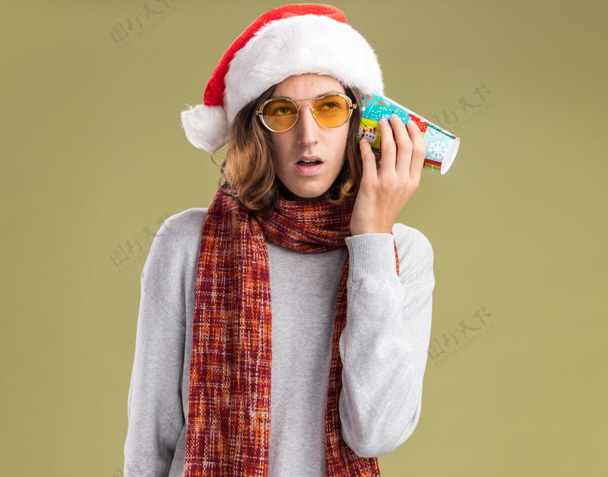 抱着快乐的年轻人戴着圣诞老人的帽子 戴着黄色的眼镜 脖子上围着暖和的围巾 耳朵上戴着五颜六色的纸杯 站在绿色的背景下显得很困惑年轻围着戴着