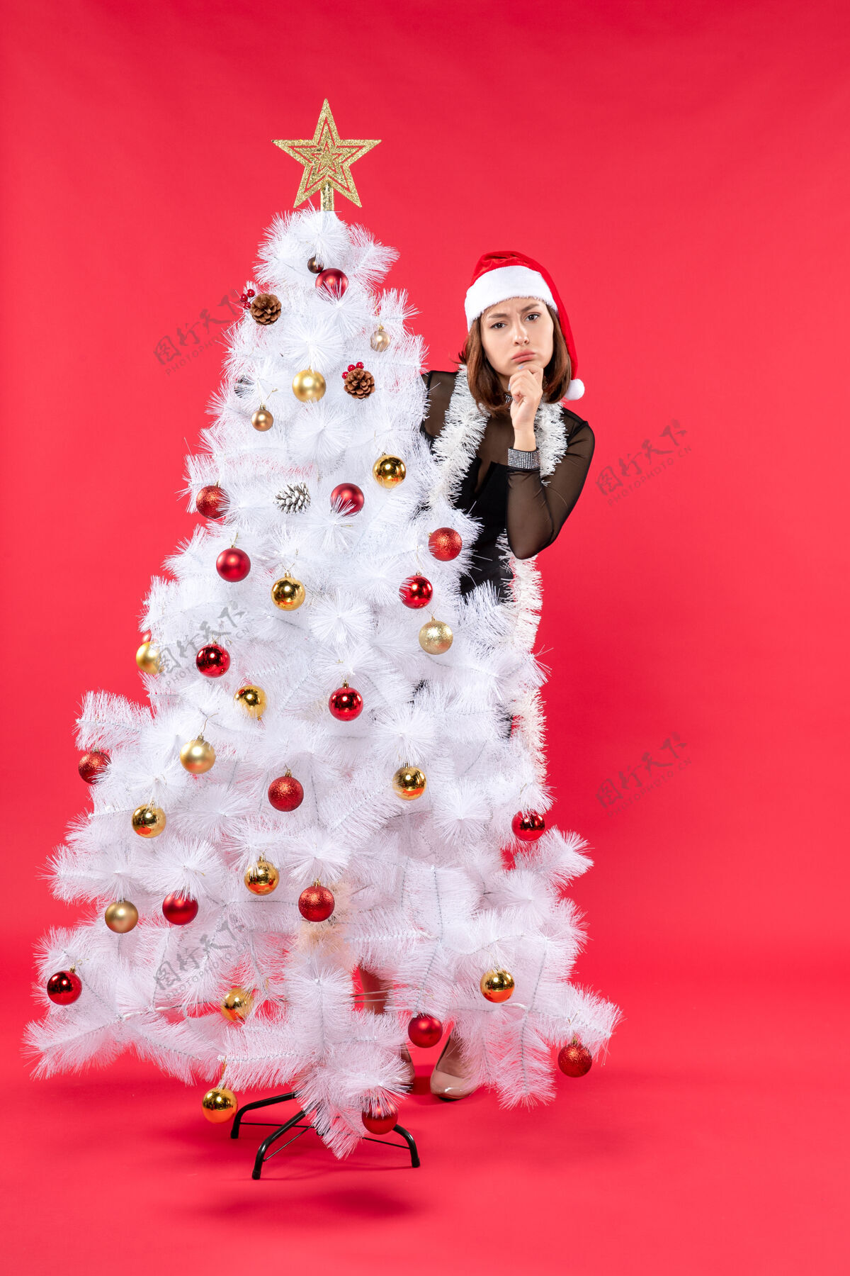 衣服圣诞树后面藏着一个穿着黑色连衣裙 戴着圣诞老人帽子的漂亮女孩 新年气氛的垂直视图新娘思念帽子