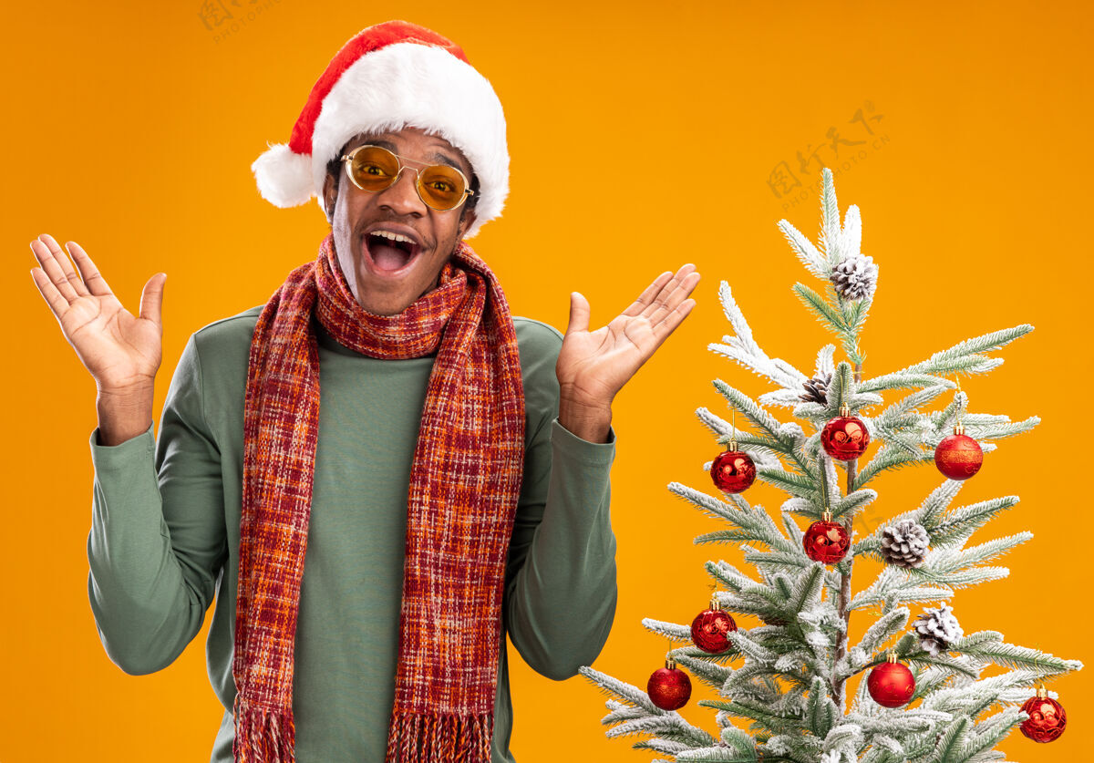 圣诞戴着圣诞帽 围着围巾的非洲裔美国人站在橙色背景下的圣诞树旁 开心地看着镜头男人周围相机