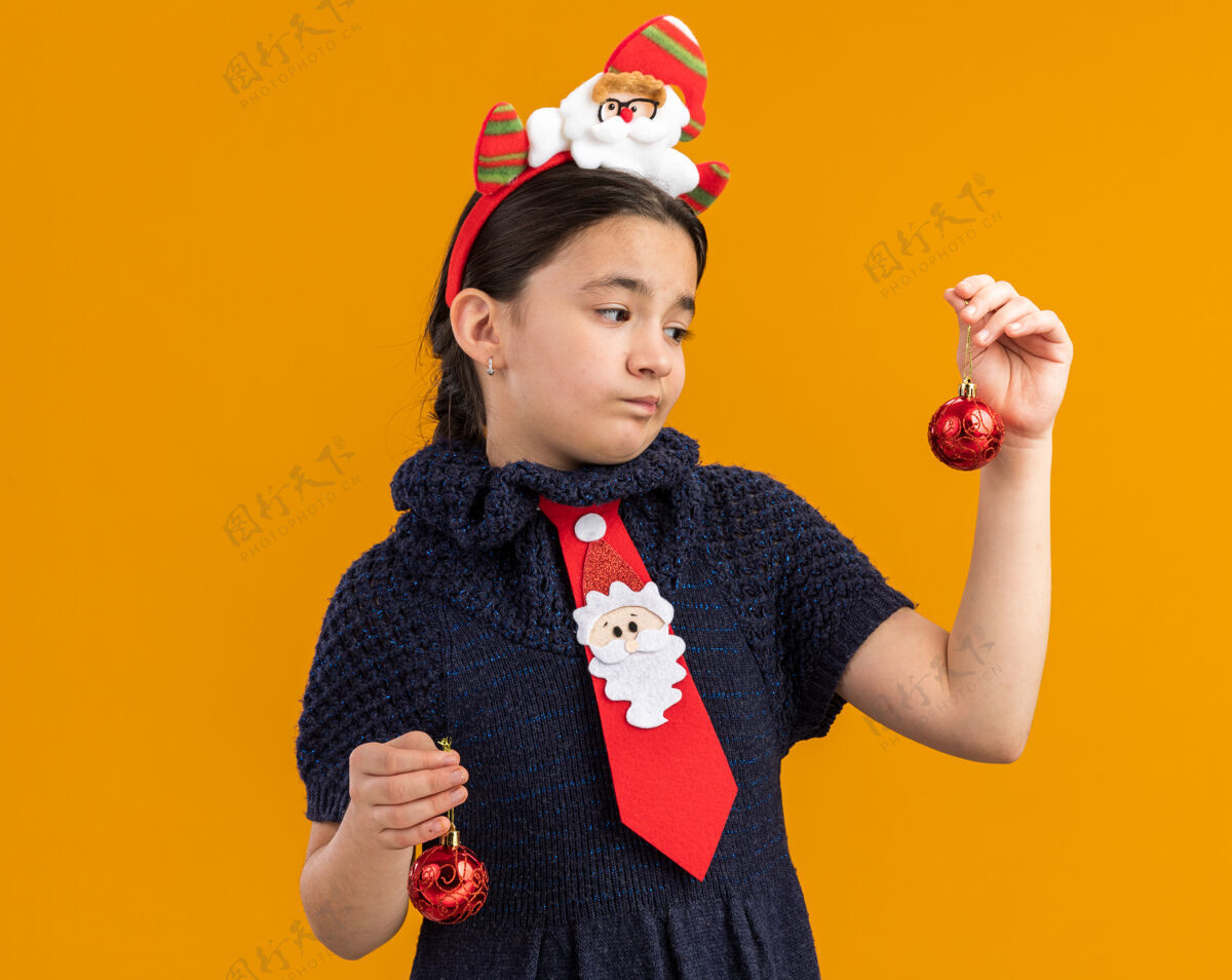 抱着穿着针织连衣裙的小女孩头上系着一条红领带 手里拿着圣诞球 神情迷茫 心中充满疑惑裙子红色搞笑
