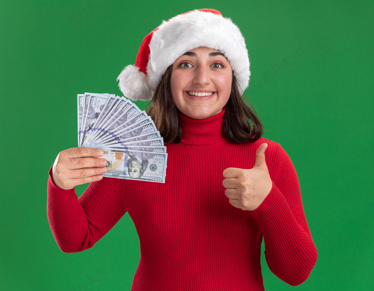 圣诞老人穿着红毛衣 戴着圣诞帽 手里拿着现金 高兴地竖起大拇指站在绿色的墙上的快乐女孩圣诞节拇指现金