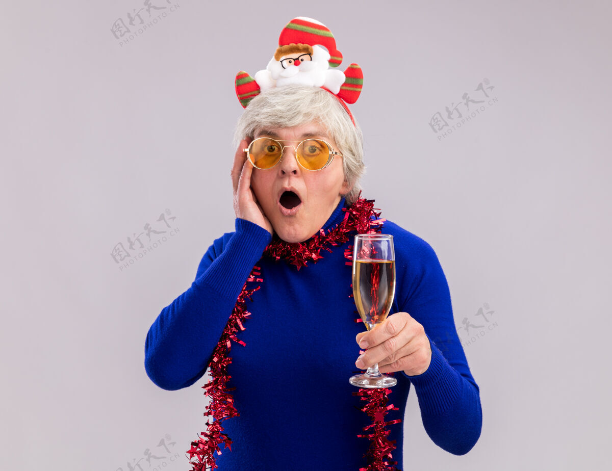 圣诞老人戴着太阳眼镜 脖子上戴着圣诞老人头带和花环的震惊的老妇人把手放在脸上 手里拿着一杯香槟圣诞快乐新年圣诞