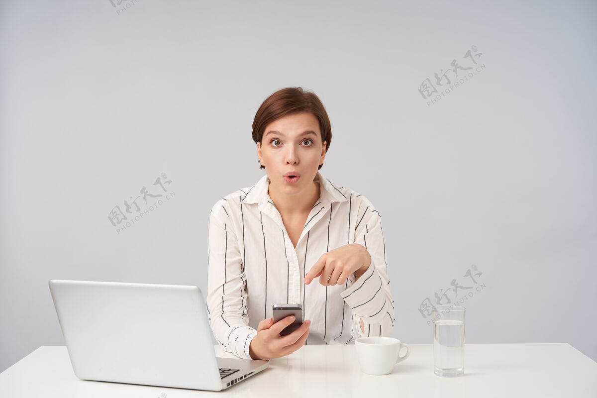 工作一位年轻可爱的黑发短发女士穿着条纹衬衫 手里拿着智能手机 圆着棕色的眼睛 坐在白色的椅子上惊奇地看着模特正式肖像