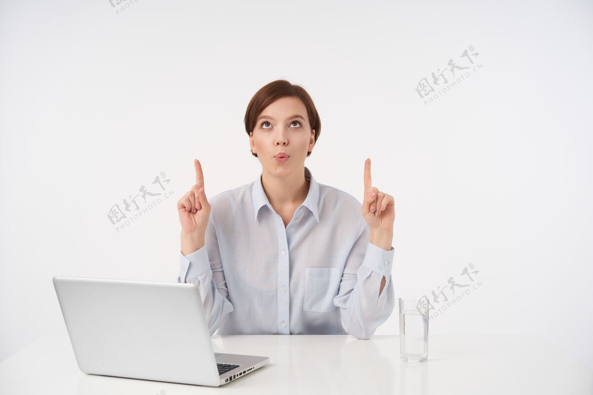 积极室内画像 年轻的棕色眼睛短发女士 自然化妆 双手放在台面上 食指向上 穿着蓝色衬衫 坐在白色椅子上姿势自然坐着