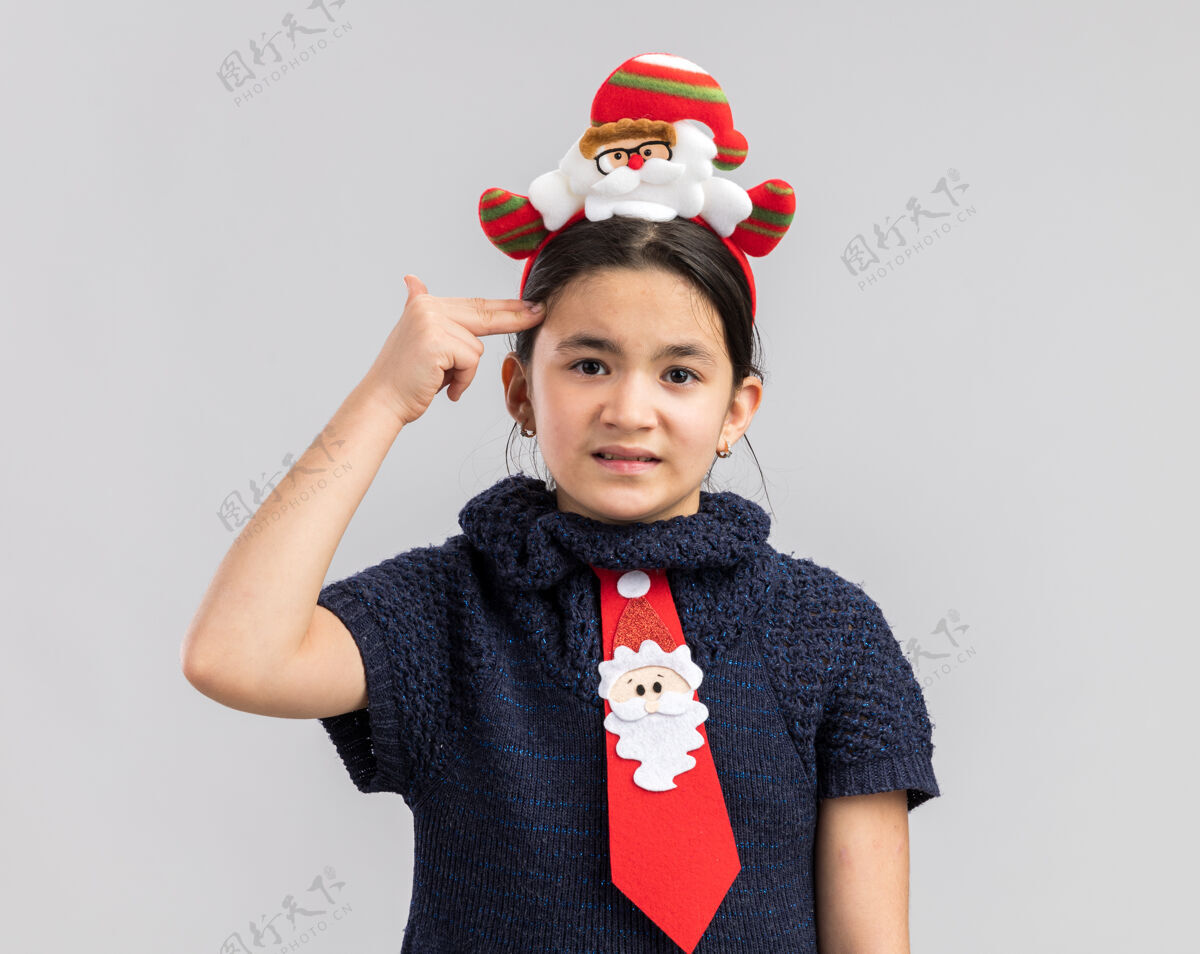 小穿着针织连衣裙的小女孩头上戴着一条红色领带 上面戴着一个滑稽的圣诞戒指 看上去很恼火 手指举过头顶做着手枪手势手势编织手指