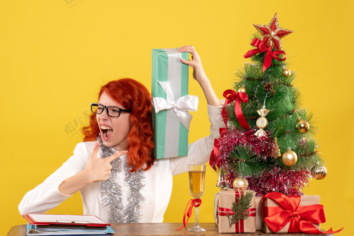 成人正面图女医生坐在黄色的桌子上 手里拿着圣诞礼物和圣诞树漂亮快乐肖像