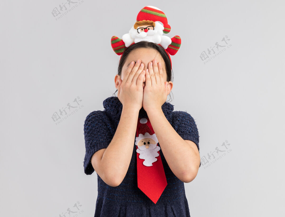 脸穿着针织连衣裙的小女孩 头上系着一条红色领带 上面戴着一个滑稽的圣诞边 手捂着脸针织女孩穿